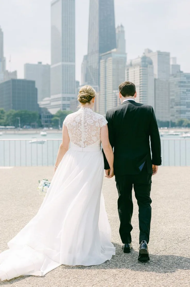 Back of bride in a high-neck lace bodice gown and organdy skirt, holding hands with her groom facing a city skyline.