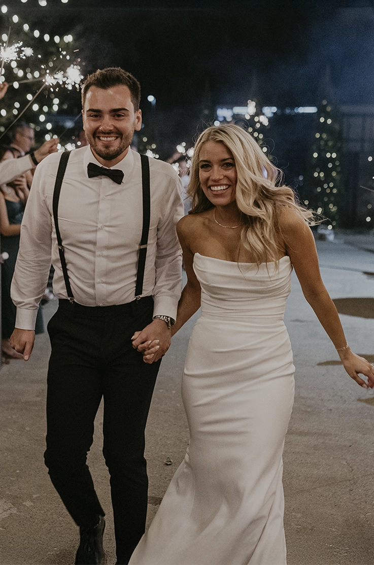 Bride in a draped crepe bodice wedding dress with a fitted skirt, walks hand in hand with her groom past sparklers at night.