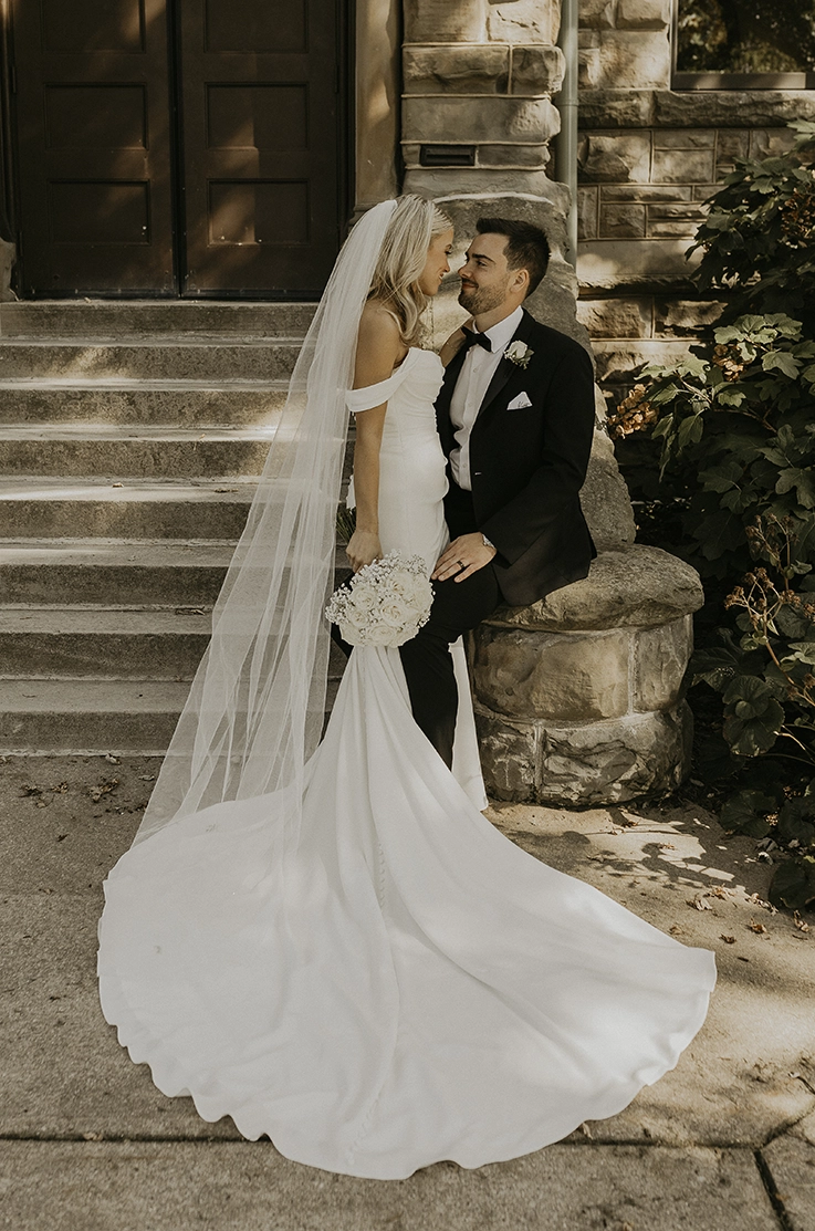 Bride in a crepe fit and flare gown with draped sleeves and a veil, poses with her groom at outdoor stone steps.