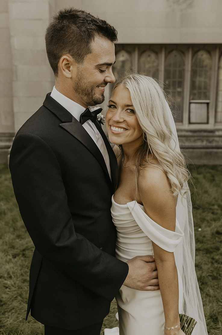 Groom embraces his bride in an off-shoulder crepe gown with draping and a veil, smiling outside the venue.