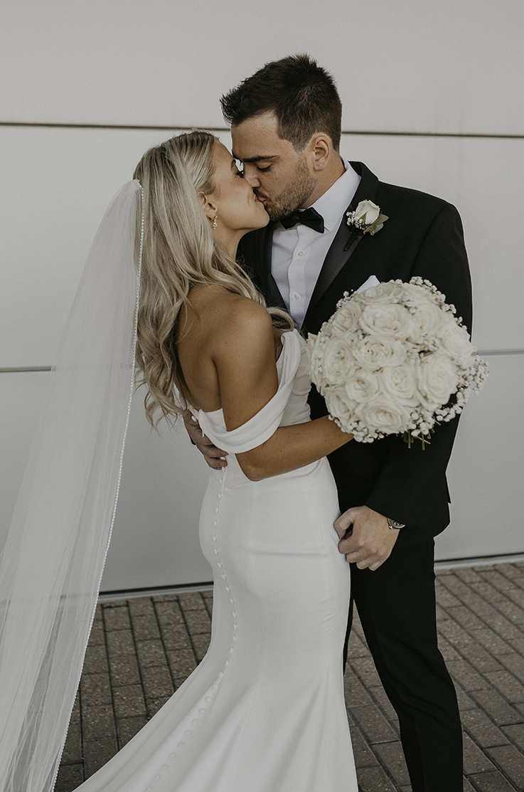 Groom kisses his bride in a crepe fit and flare gown with buttons, draped sleeves and a veil, holding a white rose bouquet.