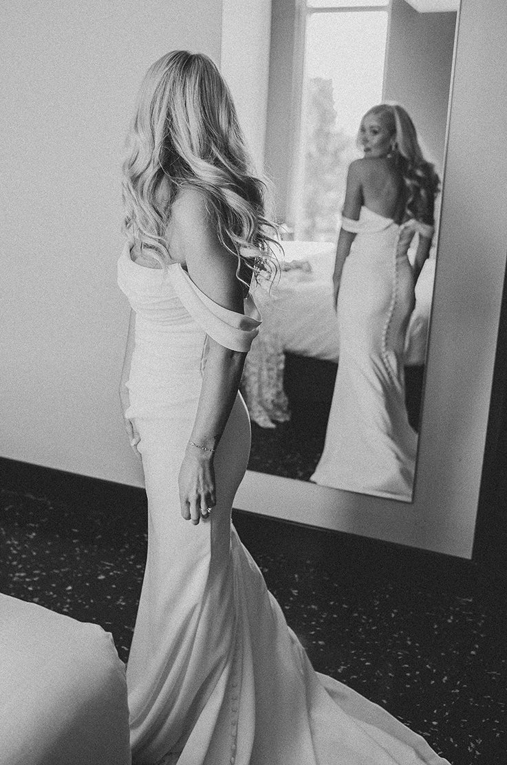 Bride looks in a mirror indoors, in a scoop neck crepe gown with draping, off-shoulder sleeves and a fit and flare skirt.