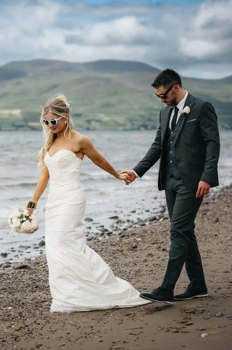 Bride in a strapless silk ruched gown with a fit and flare skirt and lace beading, holds hands with groom on a shoreline.