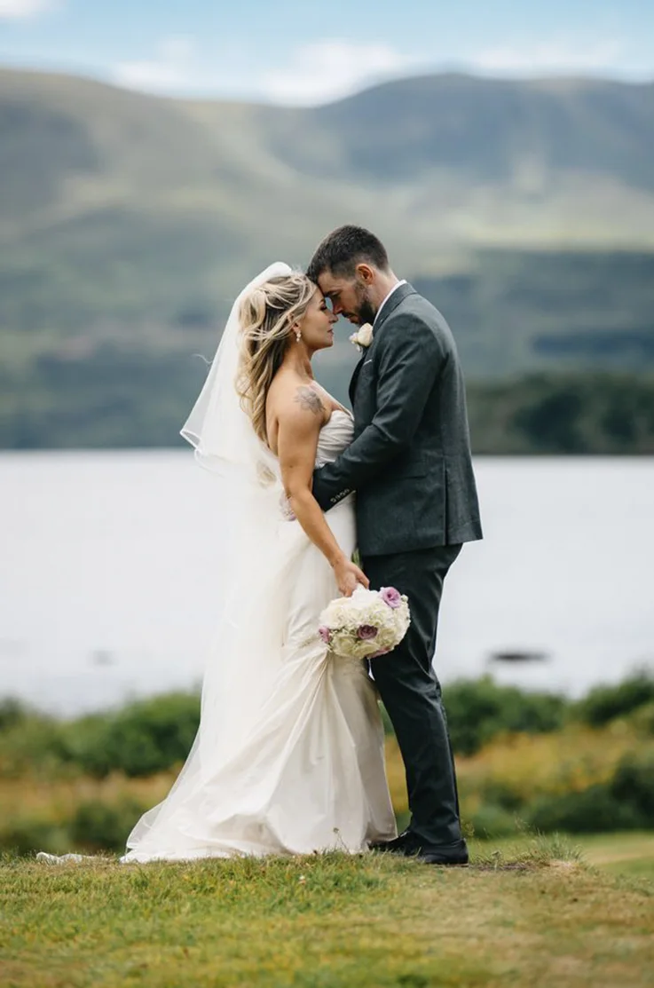 Groom embraces bride in a strapless silk gown with a pleated bodice and a veil, standing on a hill next to lake and mountains.