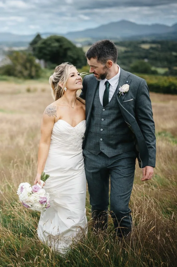 Bride in a strapless fitted silk wedding dress with lace detail, holds a bouquet and smiles at her groom in an open field. 