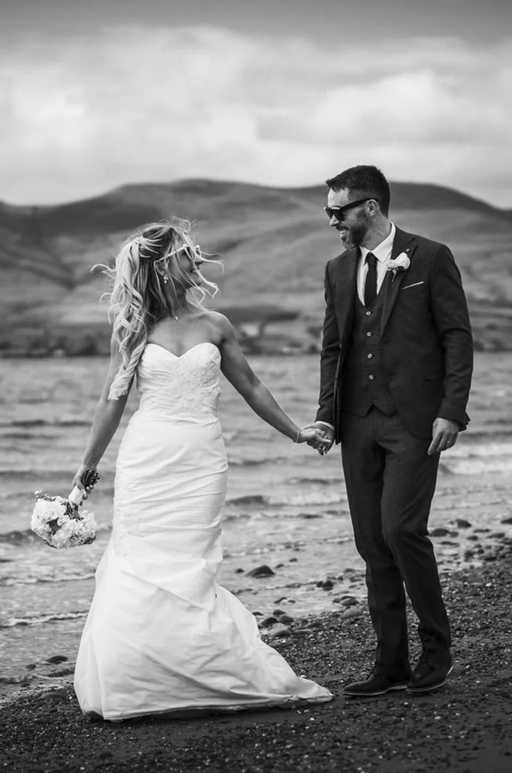 Bride in a sweetheart neck silk gown with lace details and fitted skirt, poses with groom on a lakeshore, in black and white. 
