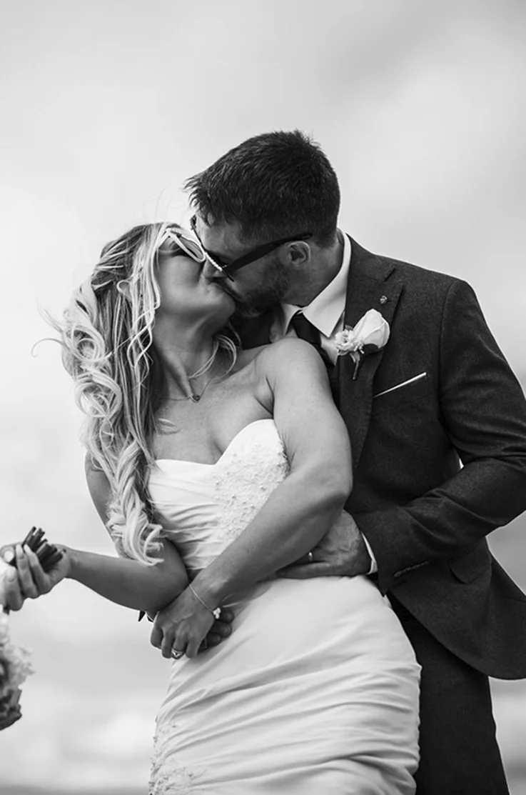 Groom holds bride in an embrace, in a sweetheart neck silk gown with beaded lace applique and they kiss in black and white. 