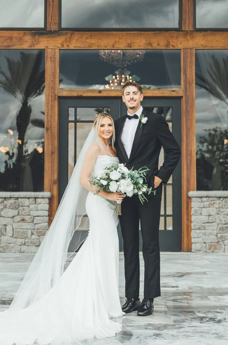 Bride in a fit and flare crepe wedding dress with draping and a veil, poses next to groom in a tux in front of venue entrance.