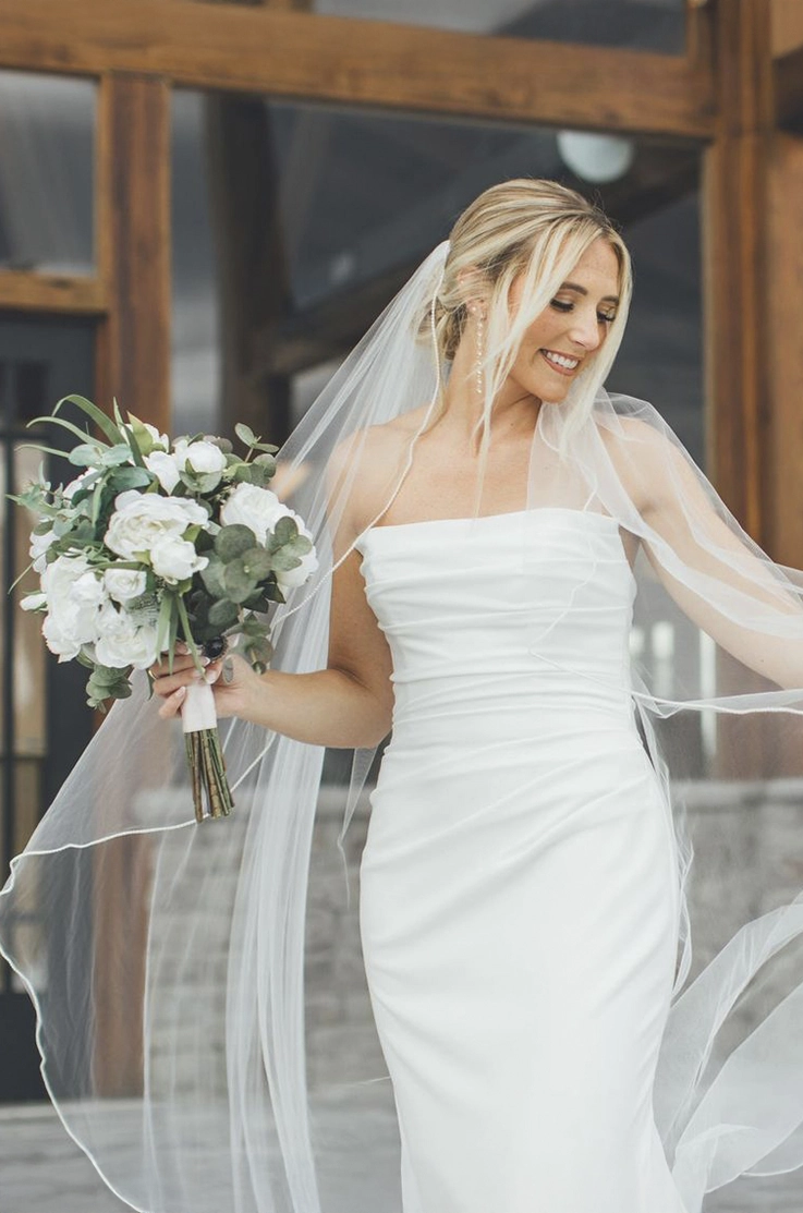 Close-up of bride smiling in a draped crepe bodice fitted gown with a flowing veil, and is holding a bouquet outdoors.
