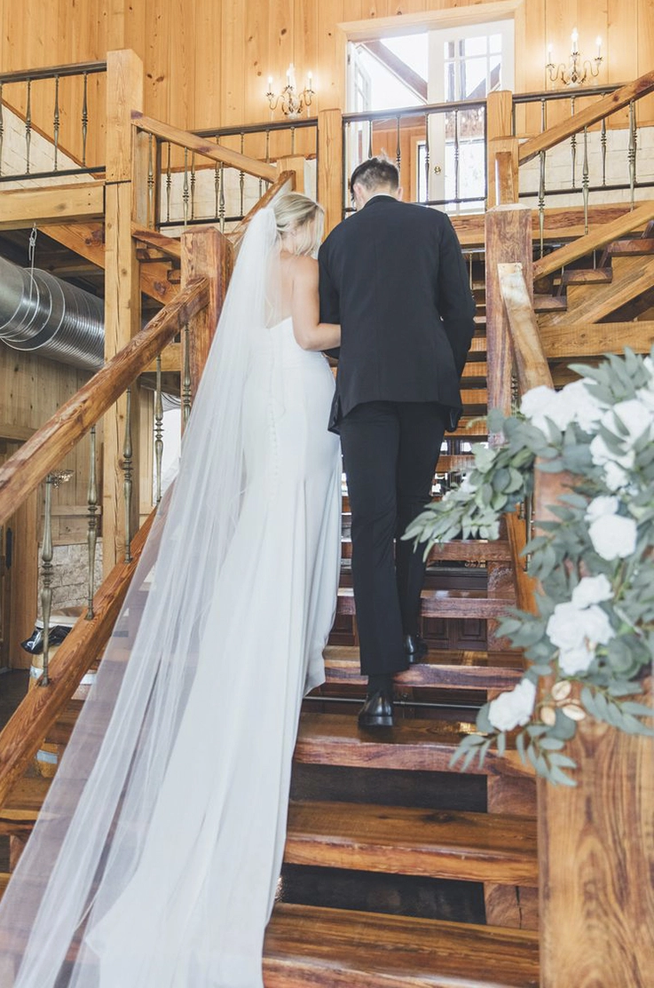 Bride in a crepe fit and flare gown with a buttoned back and a long veil, walks up a wooden staircase beside the groom.