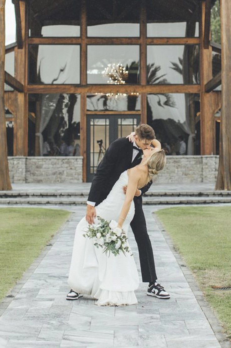 Groom dips his bride in a strapless crepe fitted gown, holding a bouquet to the side on a path leading up to the venue.