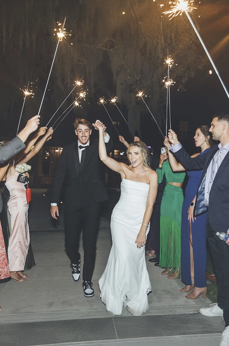 Bride in a scoop neck crepe gown with fit and flare skirt, celebrates beside groom as the guests hold up sparklers.
