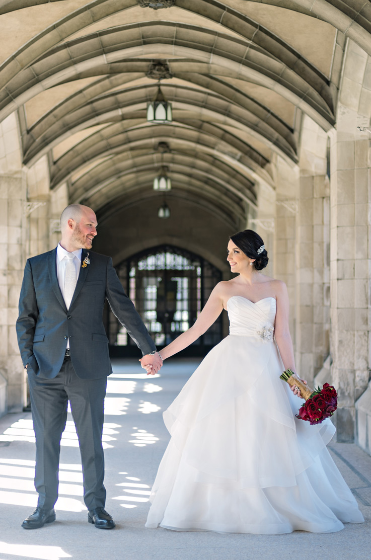 A bride in a tiered ball gown wedding dress, holding a bouquet, and a groom in a suit hold hands and look at each other in a hallways of stone archways.