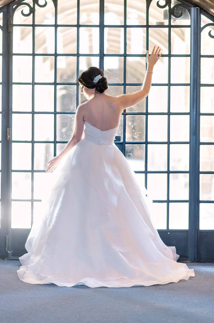 A bride in a ball gown wedding dress stands in front of a large window with decorative iron bars, facing the window with one hand up and one hand down.