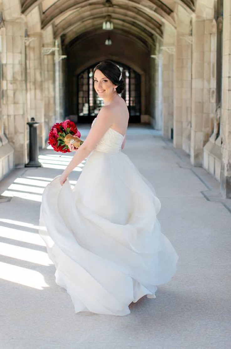 A bride in a ball gown wedding dress holds a bouquet, spins, and smiles in a hallway of stone archways.
