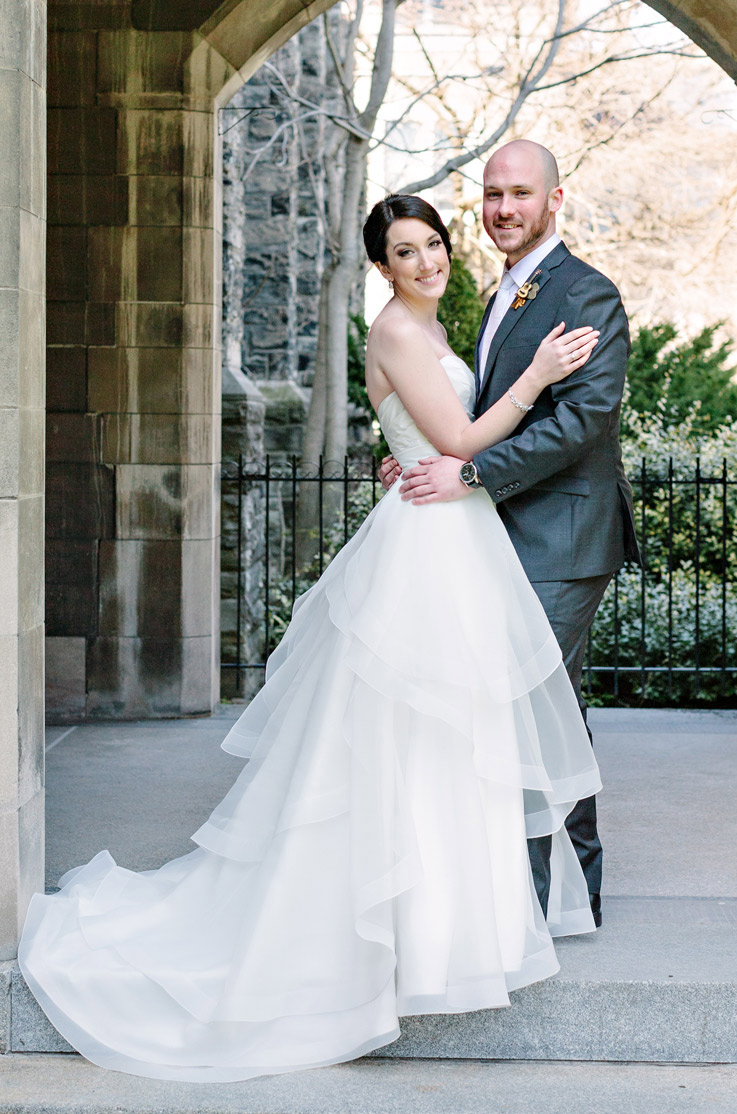 A bride in a tiered ball gown wedding dress and a groom in a suit hold each other and stand outside in a stone entranceway, with an iron gate and garden behind them. 