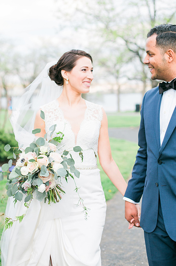 A bride in a sleeveless wedding dress and veil, holding a bouquet, looks at a groom in a suit while holding his hand and smiling, while standing outdoors.