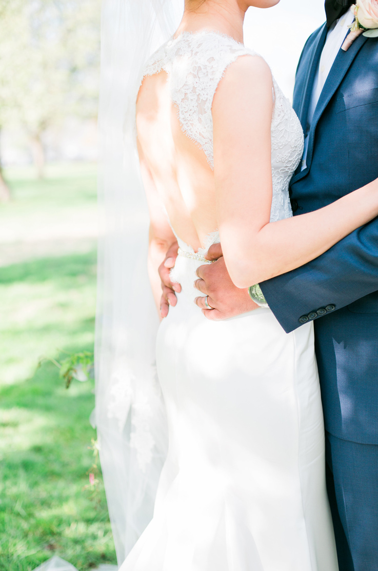 A groom in a suit holds a bride in a keyhole back wedding dress and veil close, while they stand outdoors.