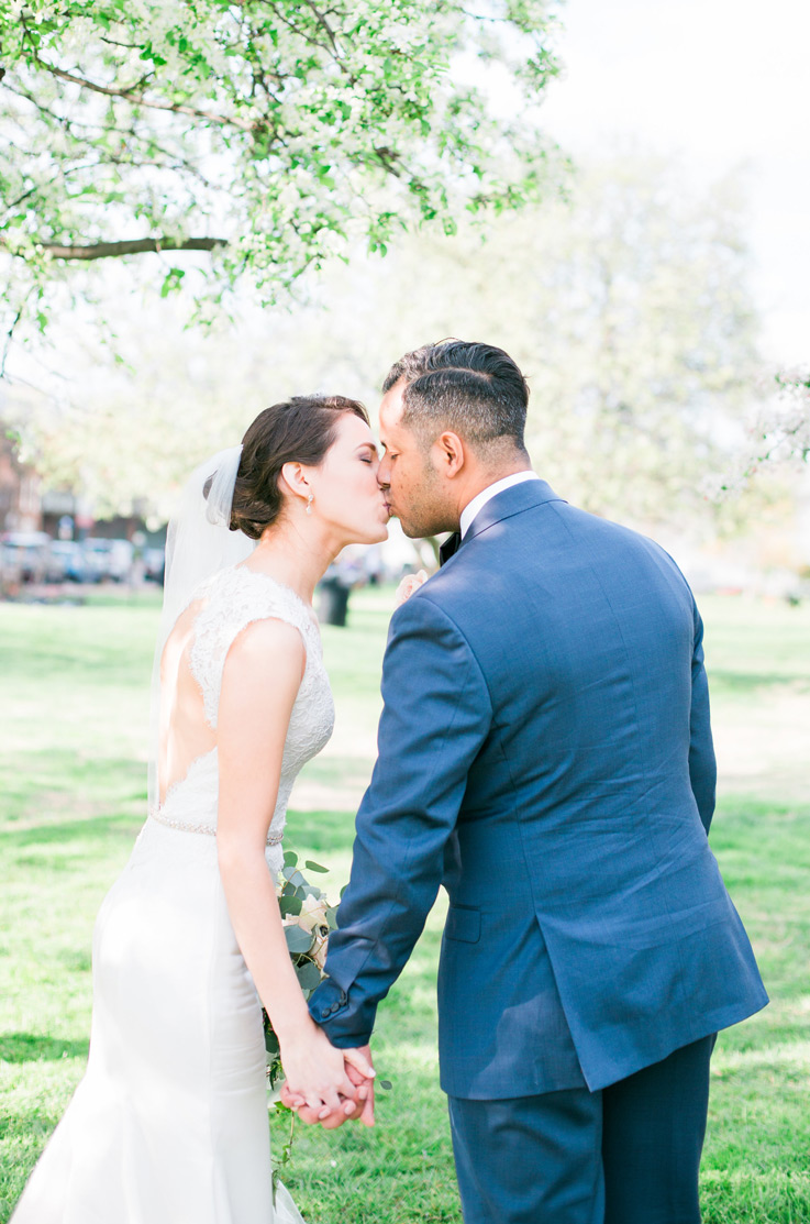 A bride in a keyhole back wedding dress and veil, and a groom in a suit hold hands and kiss while standing outdoors.