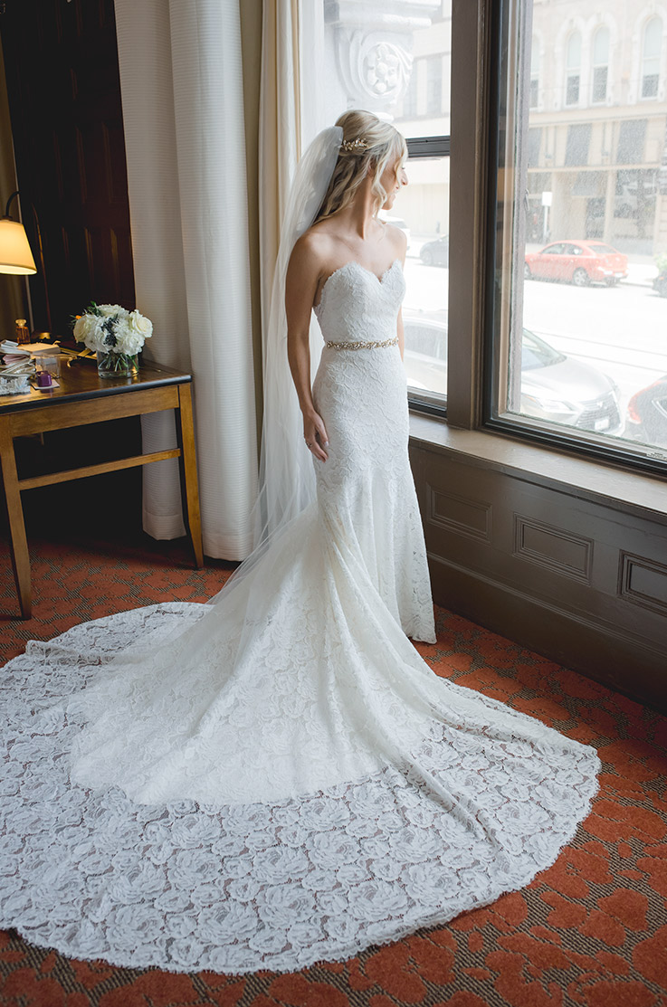 A bride in a lace wedding dress and a veil looks out a window while standing indoors, with the lace train of the dress fanned out to her side.