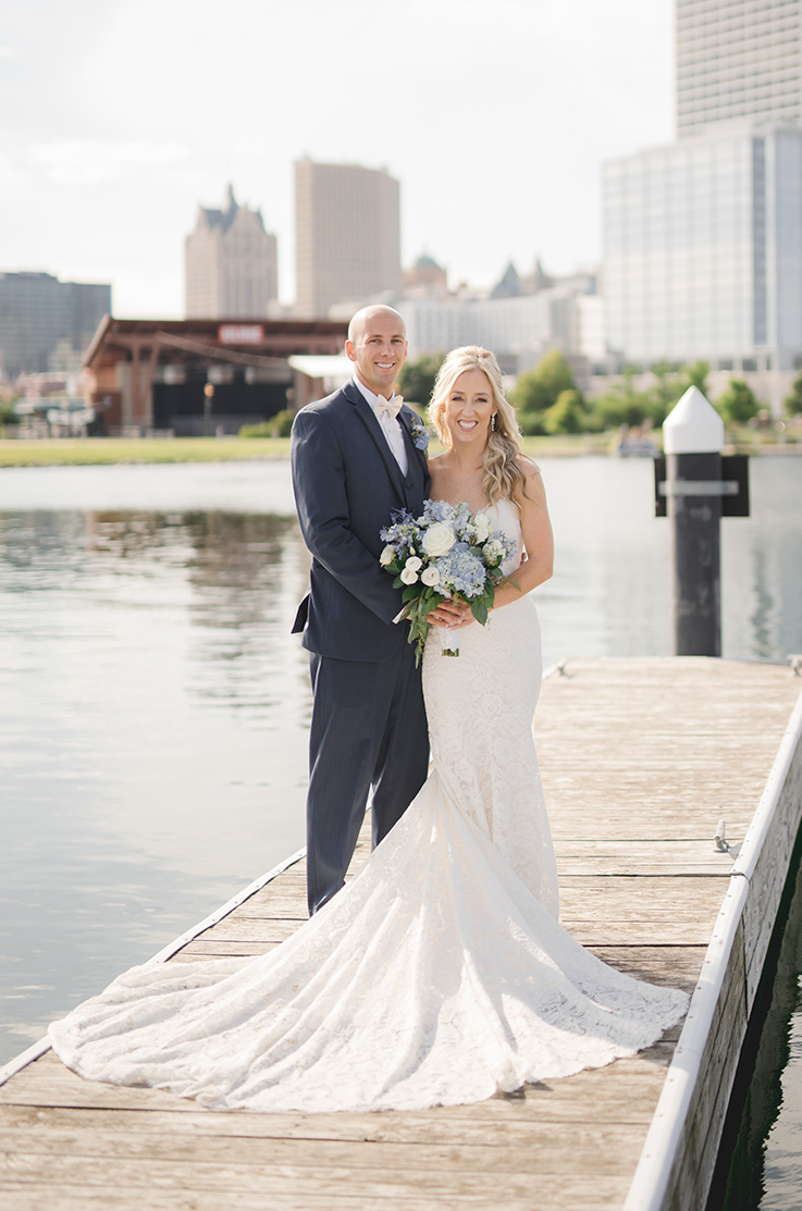 A bride in a lace wedding dress holds a bouquet and stands with a groom in a suit on a dock, with a lake behind them, and a city in the distance.