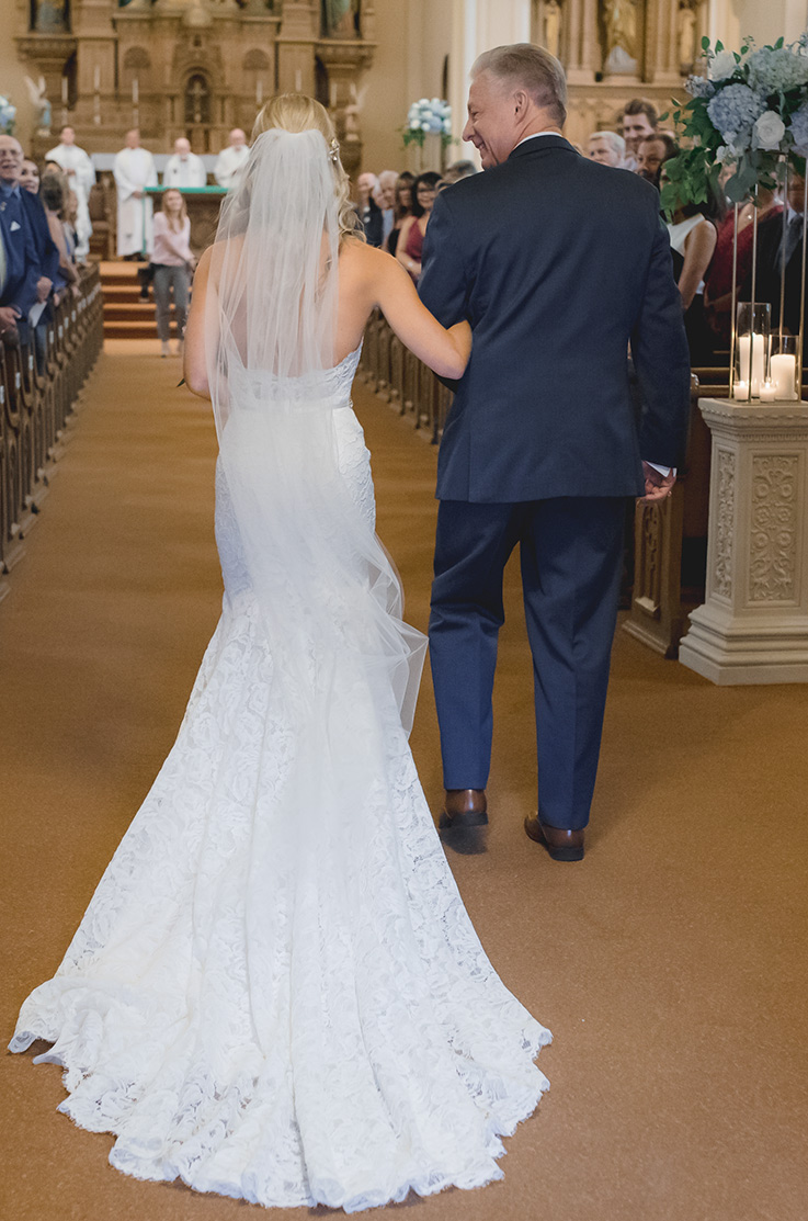 A bride in a lace wedding dress and a veil walks down the aisle towards the altar with a man in a suit, who is looking at her and smiling.