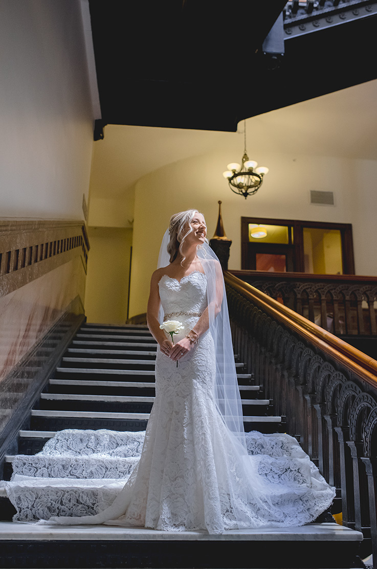 A bride in a lace wedding dress holds a rose while standing a the bottom of a staircase indoors and smiling, with her train fanned out behind her.