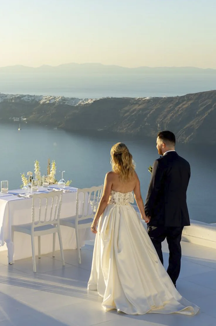Back of bride in a lace bodice gown with a full mikado skirt, holds hands with groom on a rooftop at Santorini cliffs