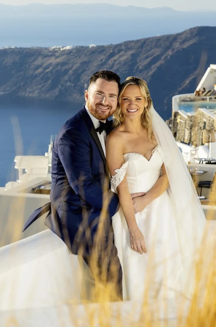 Groom poses with his bride in a mikado A-line gown with sequin lace straps and a veil on Santorini cliffside.