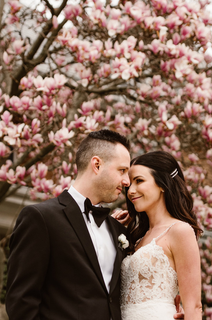 A bride in a lace wedding dress stands close to a groom in a suit, in front of a blossoming tree outdoors. 