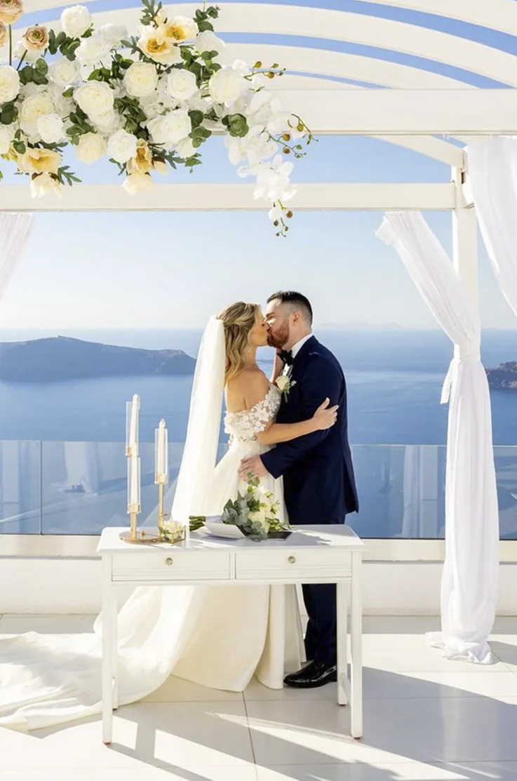 Groom kisses bride in a lace and mikado A-line gown with a veil, on a rooftop terrace at altar with floral decor at the sea. Groom kisses bride in a lace and mikado A-line gown with a veil, on a rooftop terrace at altar with floral decor at the sea. 
