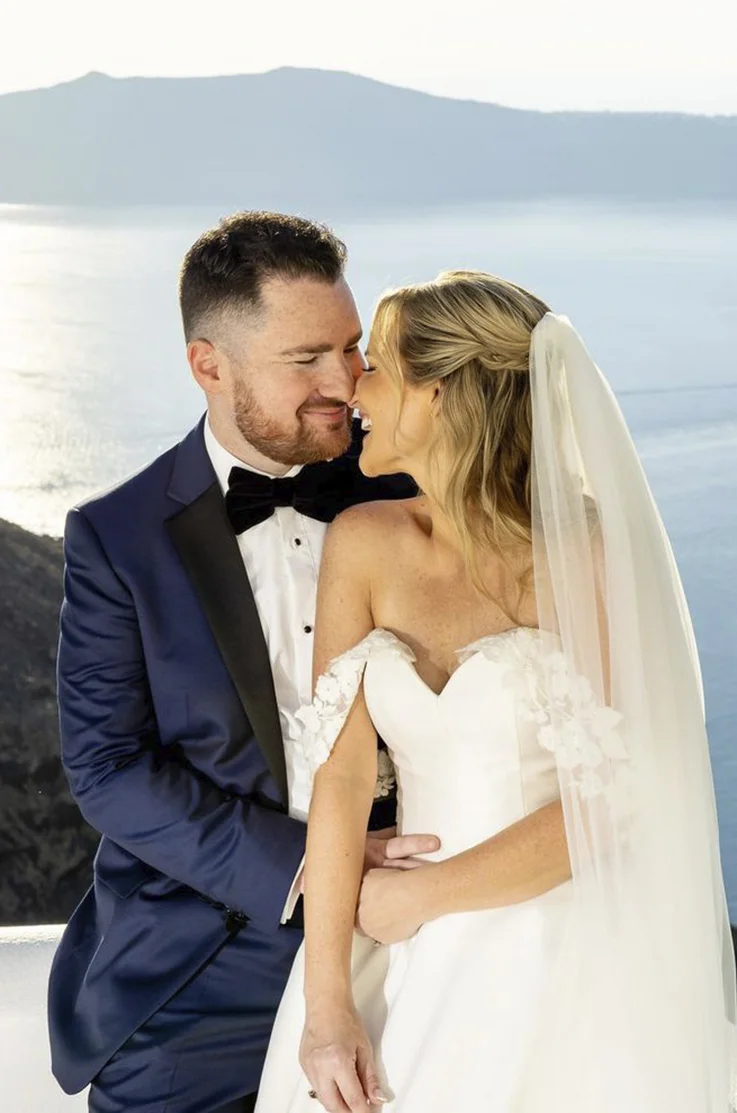 Close-up of groom in a blue suit embracing his bride in a mikado bodice wedding dress with sequin lace, overlooking the sea.