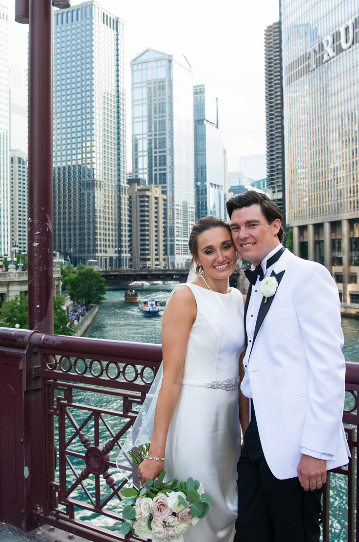 A bride in an embellished wedding dress holding a bouquet, and a groom in a contrasting suit smile and stand on a bridge in front of a river, with skyscrapers in the background.
