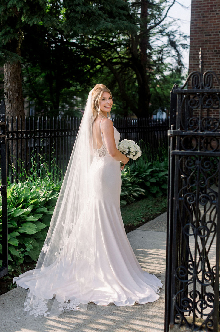 A bride in a sleeveless wedding dress and a veil looks over her shoulder and smiles, while holding a bouquet, on a path with bushes and iron fences around it.