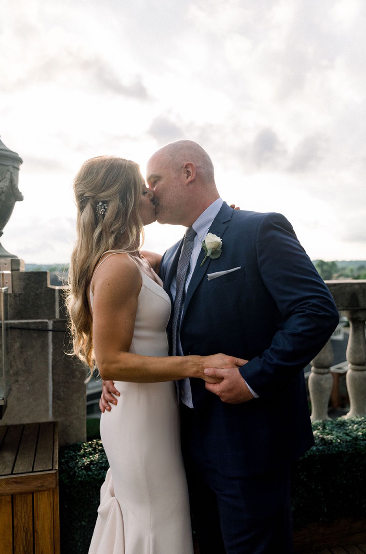 A bride in a sleeveless wedding dress and a groom in a suit kiss while standing outside, with stone rails behind them.