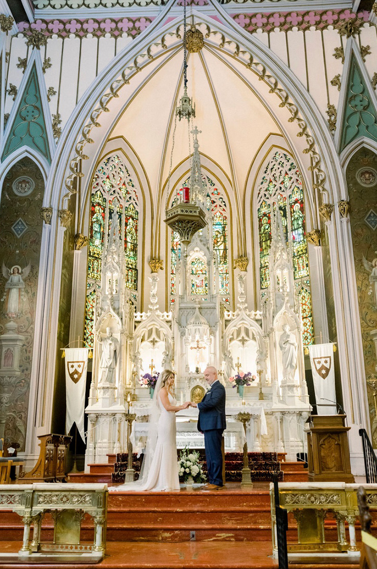 A bride in a fitted wedding dress and a veil, and a groom in a suit, hold hands and face each other at the altar of a church, in front of statues and stained glass.