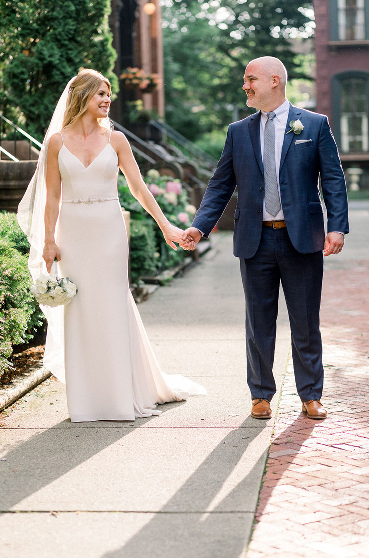A bride in a V-neckline wedding dress and veil, and a groom in a suit hold hands and look at each other and smile, while standing on a sidewalk with buildings and hedges behind them.