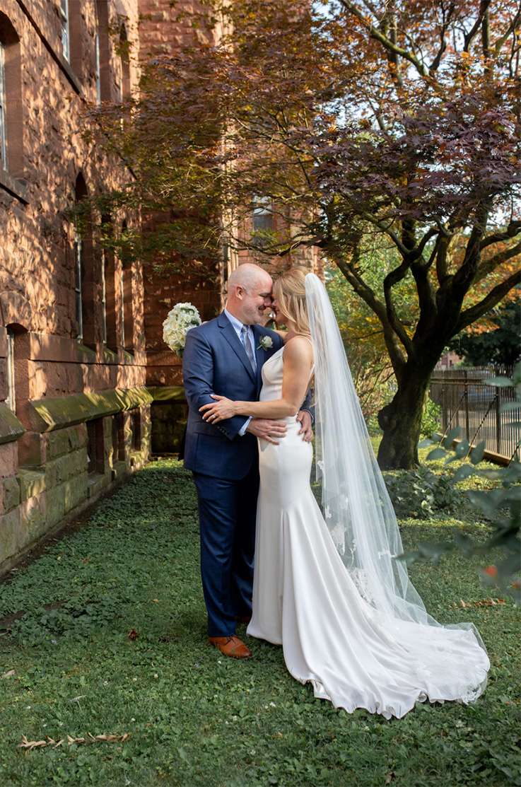 A groom in a suit holds a bride in a sleeveless wedding dress close, both looking at each other and smiling. They are standing in front of a brick building, in front of a tree.
