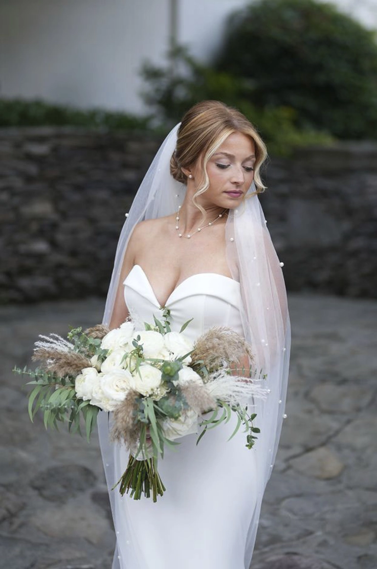 Bride poses outdoors in a strapless fitted crepe wedding dress with a pearl veil, and is holding a bouquet in a courtyard.