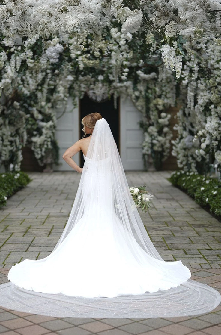 Back of bride in a crepe fit and flare gown with a pearl veil, holding a bouquet to the side beneath a floral archway.