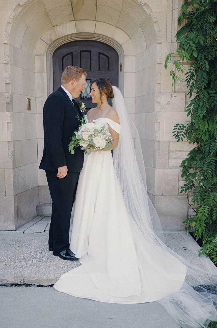 Bride in a satin faille wedding gown with a veil and off-shoulder sleeves, is facing her groom at the venue entrance.
