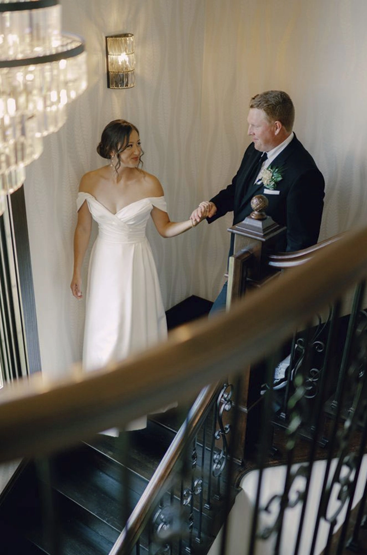 Bride in an off-shoulder satin faille gown with a draped bodice, holds hands with groom on a stairs under a chandelier.