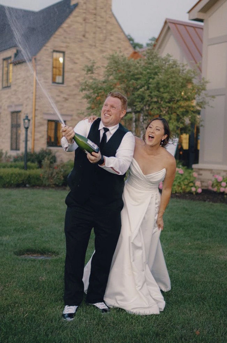 Bride in a strapless satin wedding dress with draping, pops open champagne celebrating with the groom outside the venue.
