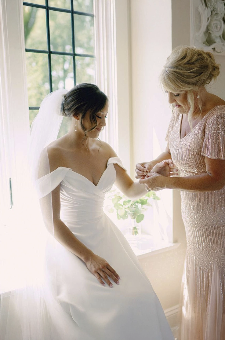 Close-up of bride getting ready by a bright open window, in a draped satin gown with off-shoulder sleeves and a long veil.