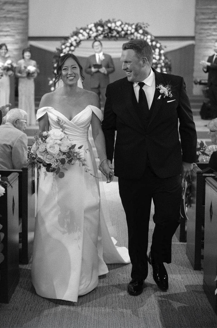 Bride in a draped satin faille gown with sleeves, smiles holding a bouquet, as she walks beside groom down the aisle