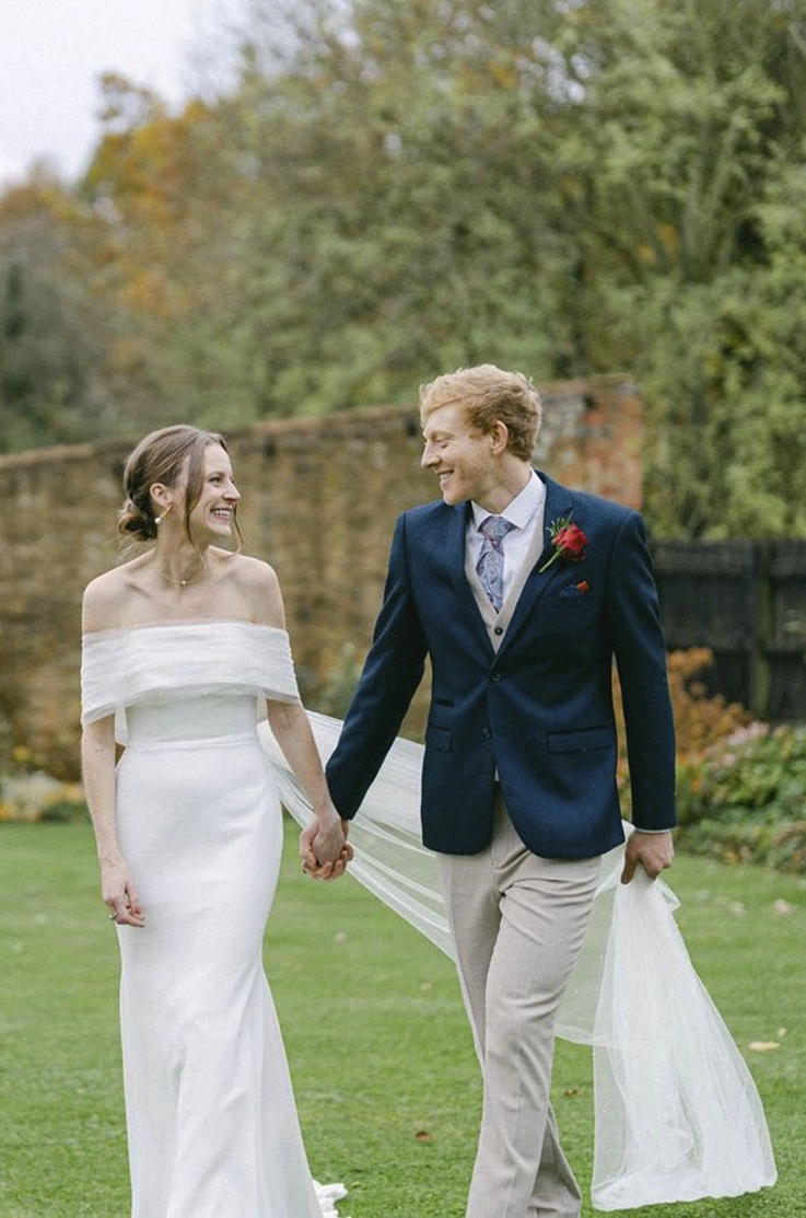Bride in a crepe potrait collar gown with fit and flare skirt and a veil, walking alongside her groom in lush gardens.