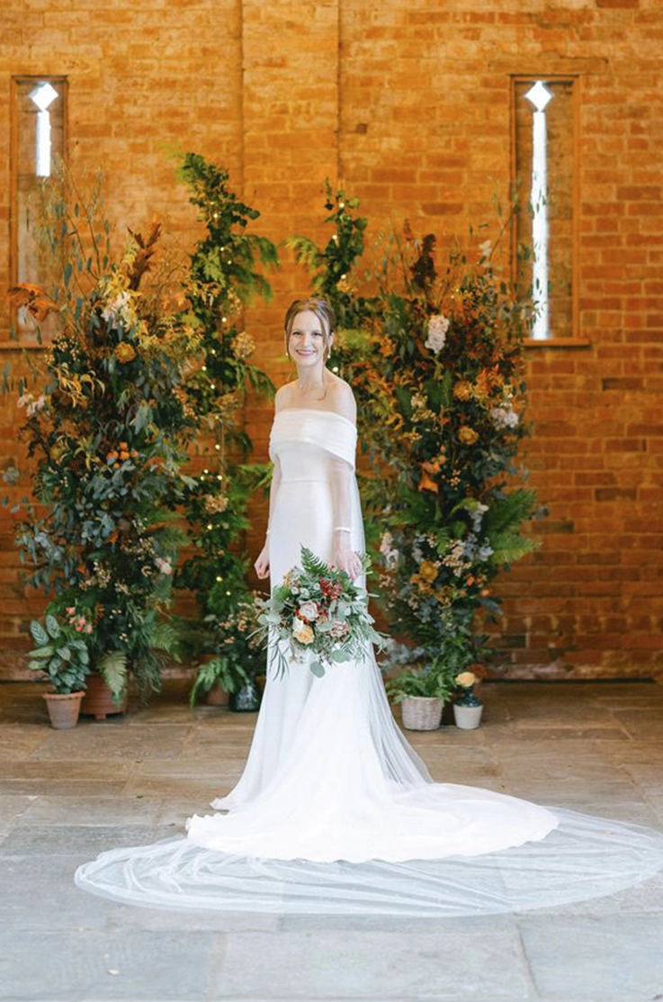 Bride in a strapless crepe fit and flare gown with a portrait collar and veil, is holding a bouquet by a brick wall with flowers. 
