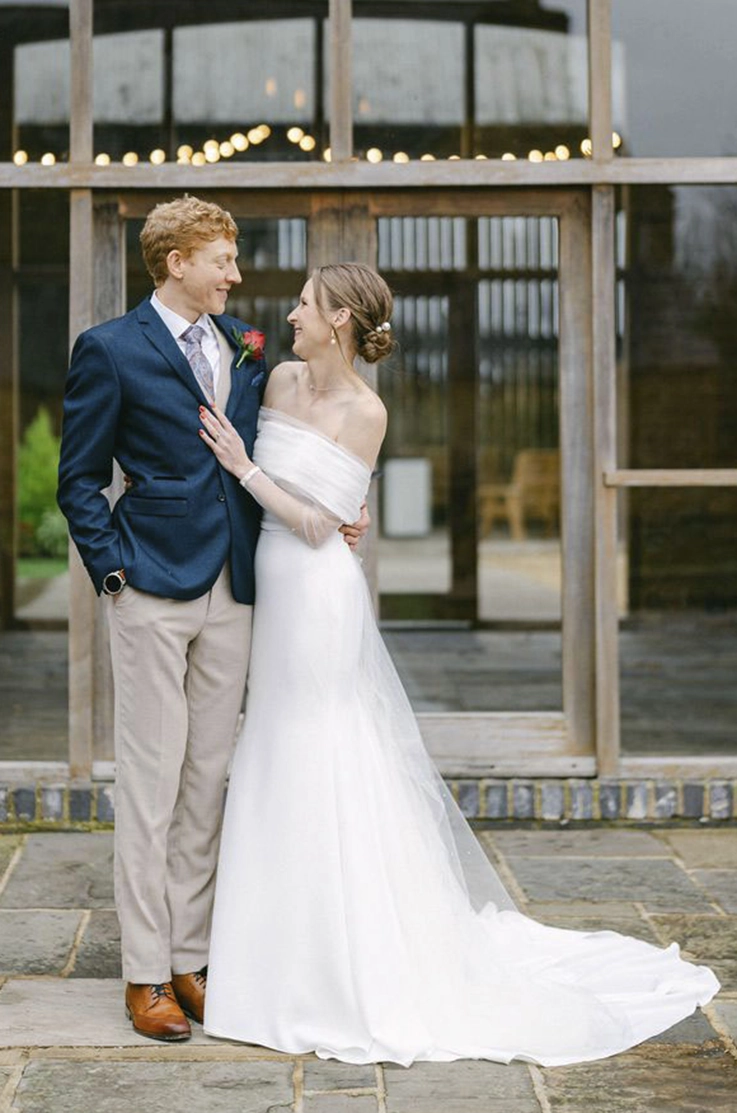Bride in an off-shoulder crepe fit and flare wedding dress with portrait collar, poses with her groom, at venue entrance.