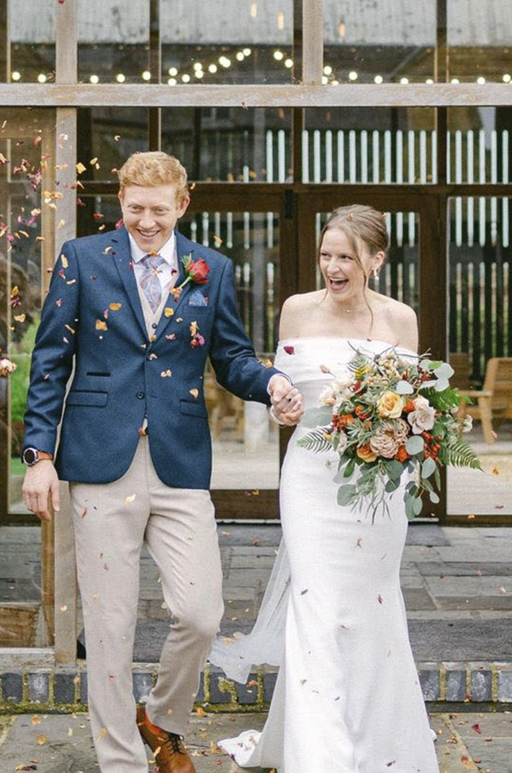 Bride in an off-shoulder fitted crepe gown with a veil, walks hand in hand with groom, and holds a bouquet as they exit.