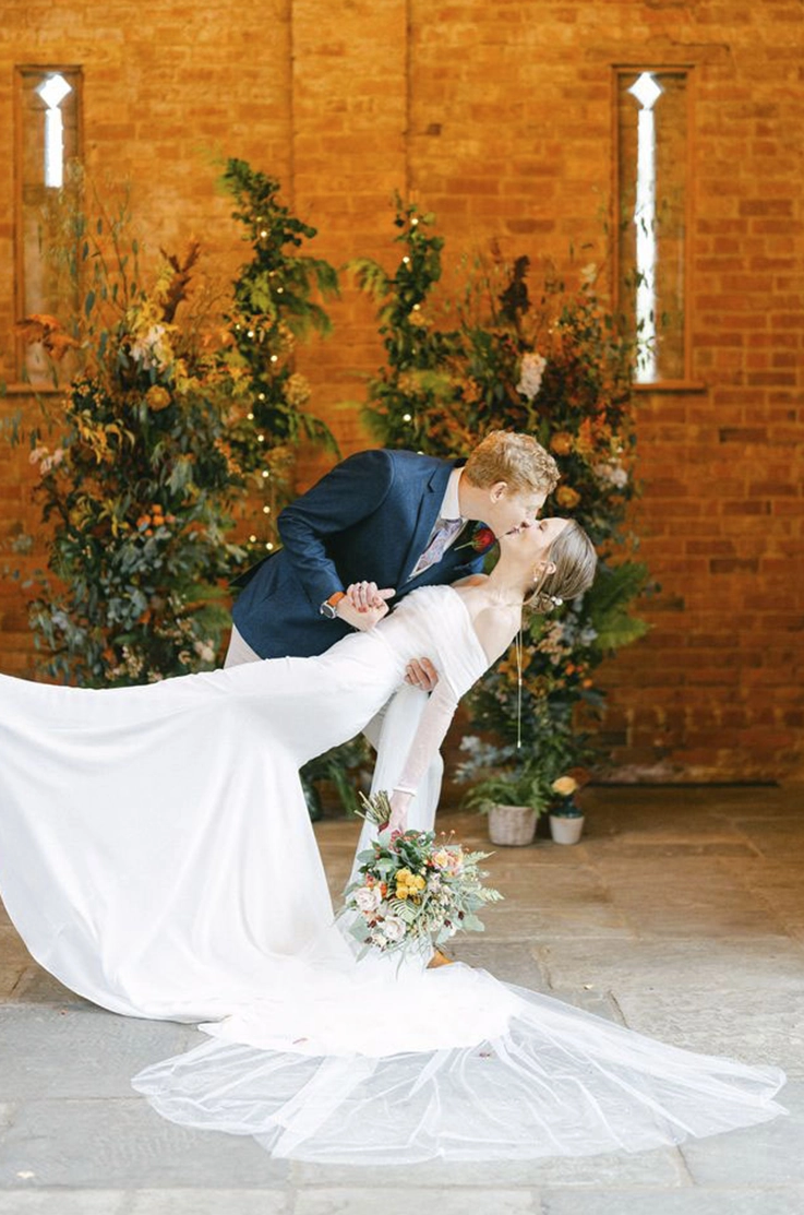 Groom dips his bride in an off-shoulder fitted crepe gown with a veil, holding a bouquet at the altar with flower arches.